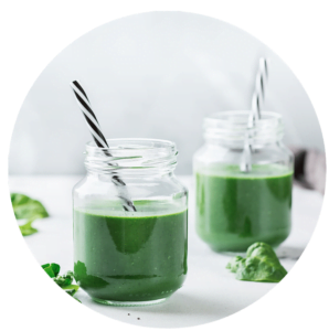 Two glass jars filled with green smoothie, each with a black-and-white striped straw, sit on a white surface with spinach leaves nearby—perfect inspiration for your next smoothie recipes.