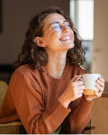 A person with curly hair and glasses is smiling while holding a white mug, wearing a brown sweater, and sitting indoors near a window, feeling refreshed after joining the Food Peace Reset Programme.