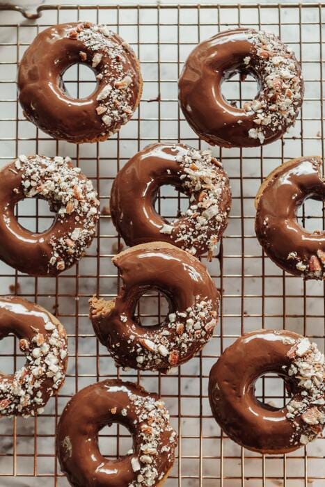 Nine chocolate-glazed donuts, some topped with crushed nuts, are arranged on a cooling rack—a tempting treat that could be enjoyed as part of your Food Peace Reset Programme; one donut has a bite taken out of it.
